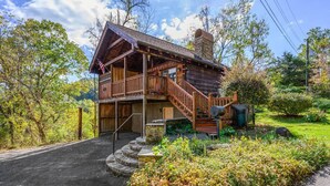 A two-story log cabin with a wooden staircase leading to the upper level, surrounded by lush greenery and a clear sky. - Location  Cute Studio Log Cabin Hot Tub Fireplace (Pigeon Forge)