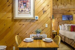 A wooden-paneled room with a dining table set for two, a bed with pillows, and a framed picture on the wall. - Location  Cute Studio Log Cabin Hot Tub Fireplace (Pigeon Forge)