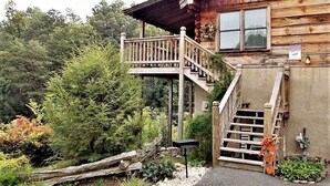 En træhytte med en trætrap, der fører op til en balkon, omgivet af frodig vegetation.