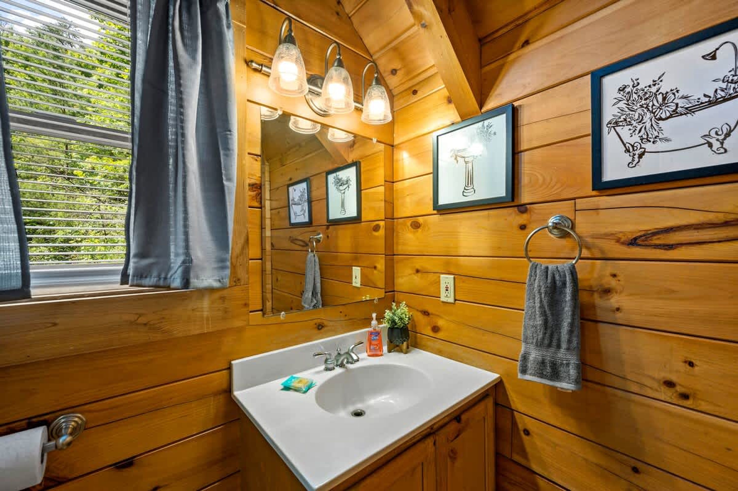 A wooden cabin bathroom with a white sink, hanging lights, framed artwork, and a window with curtains.