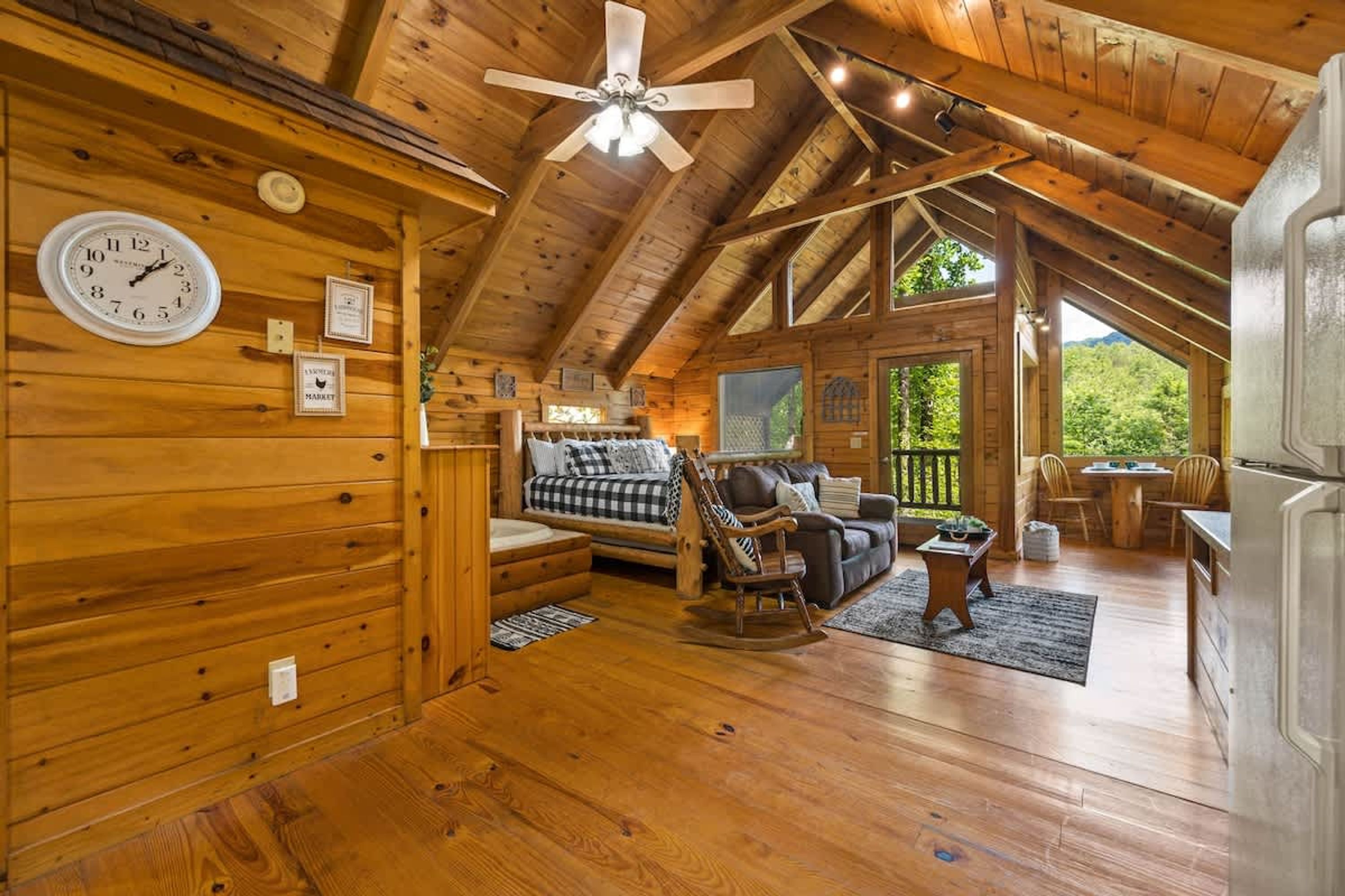 A cozy log cabin interior with a wooden ceiling, a ceiling fan, a sofa, a rocking chair, and a dining area.