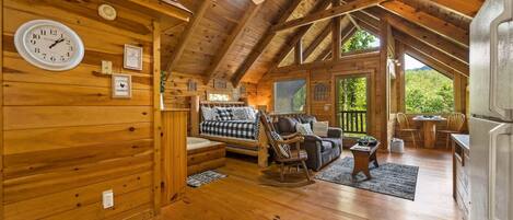 A cozy log cabin interior with a wooden ceiling, a ceiling fan, a sofa, a rocking chair, and a dining area.