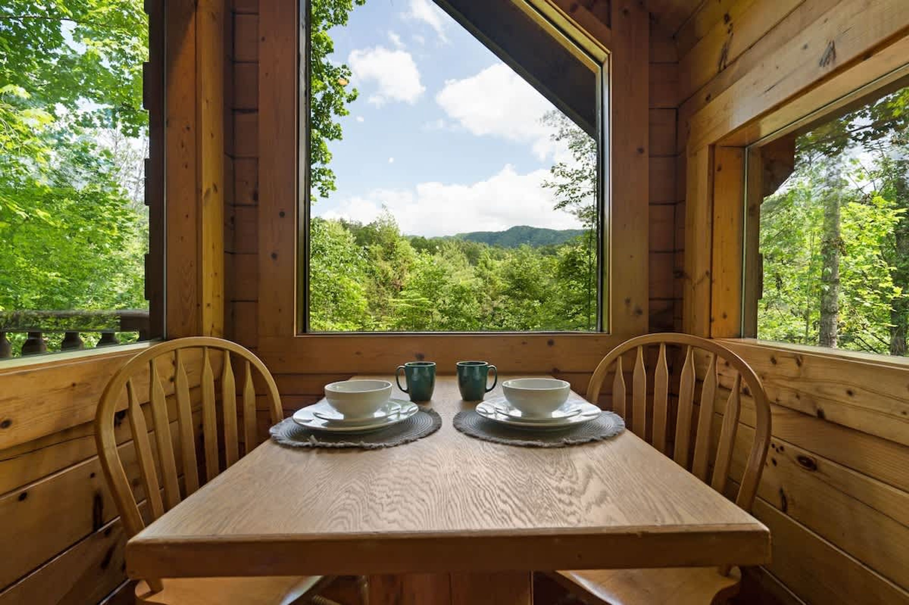 Une table à manger en bois, accompagnée de deux chaises, dressée pour deux personnes, et donnant sur une forêt grâce à une grande fenêtre.