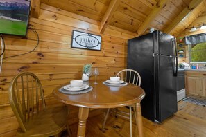 Ein Holzblockhaus-Interieur mit einem Esstisch, gedeckt für zwei Personen, einem schwarzen Kühlschrank und einem Schild mit der Aufschrift 'gather'.