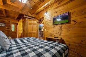 A cozy wooden cabin room with a checkered bedspread, a mounted television, and a visible door leading to another room. - Nice View  Log Cabin Fireplace Hot Tub Pool Table (Sevierville)