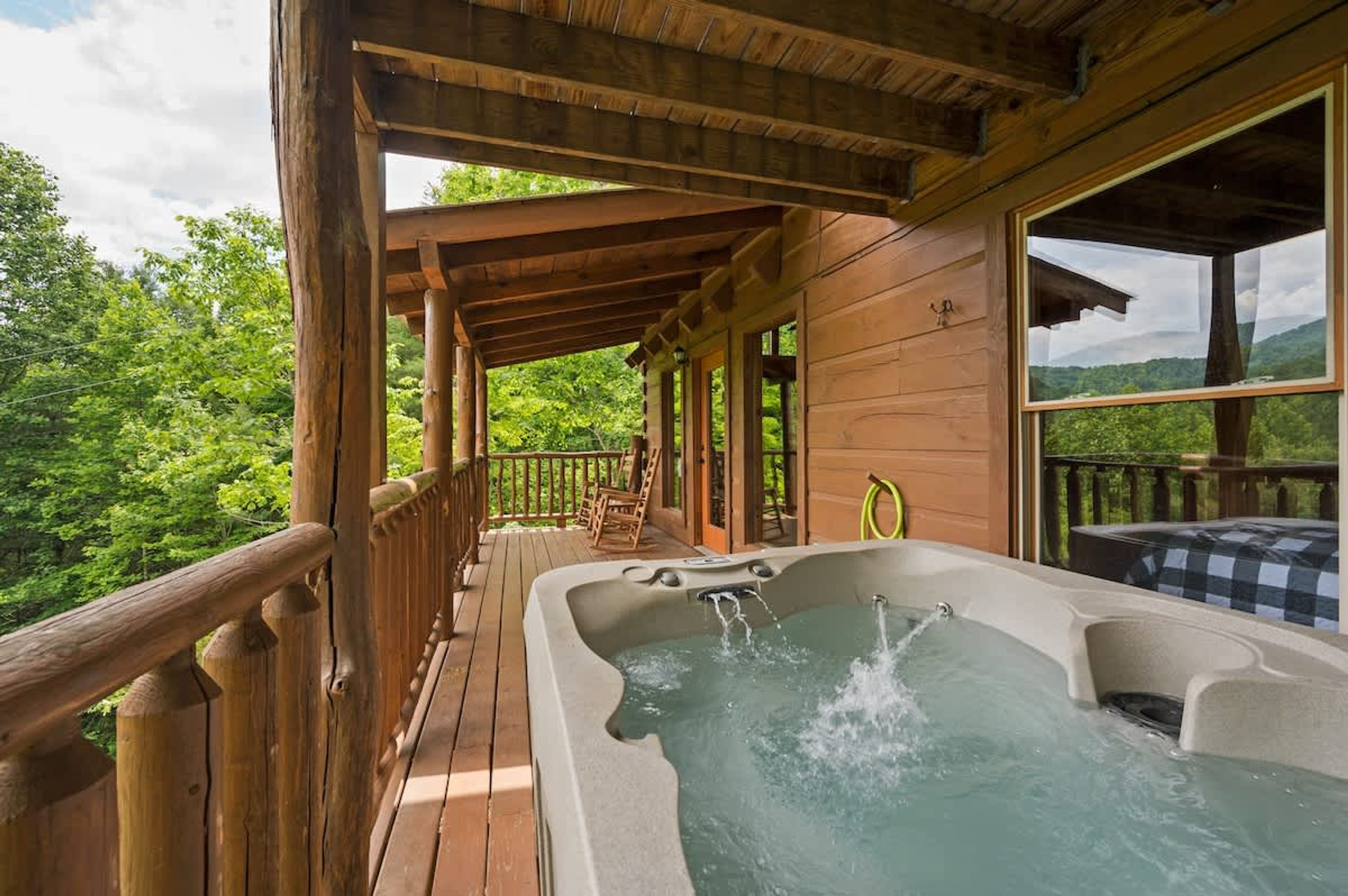 Una terraza de madera con un jacuzzi, barandas de madera y vistas a un paisaje boscoso.