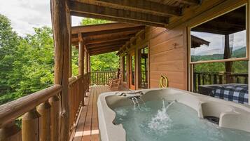 Una terraza de madera con un jacuzzi, barandas de madera y vistas a un paisaje boscoso.