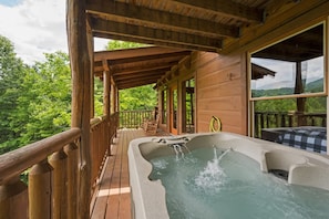 Una terraza de madera con un jacuzzi, barandas de madera y vistas a un paisaje boscoso.
