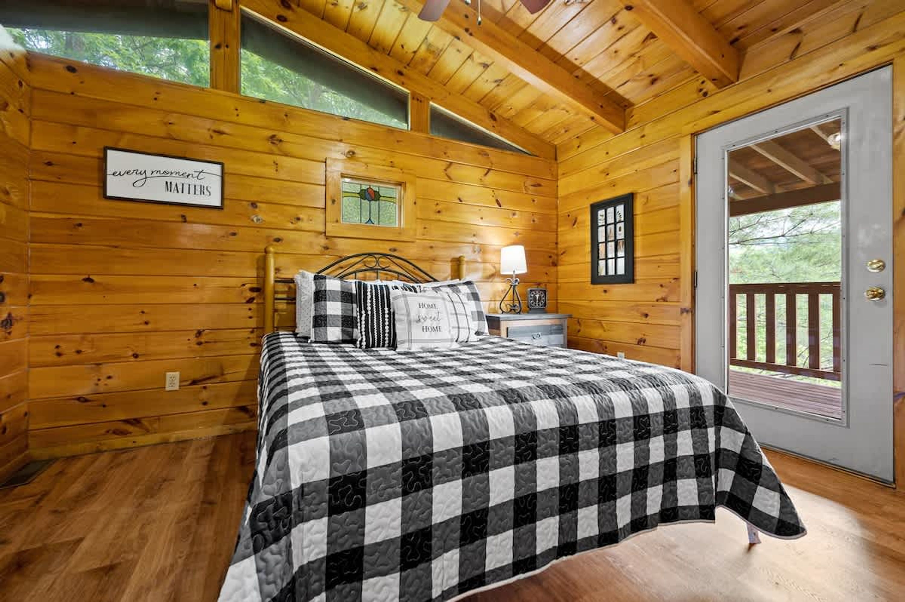 Acogedor dormitorio de cabaña de madera con colcha de cuadros, una mesita de noche con lámpara, y un letrero que dice 'cada momento cuenta'.