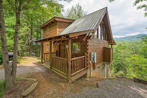 Una cabaña de madera con un porche y techo inclinado, rodeada de árboles y un paisaje montañoso.