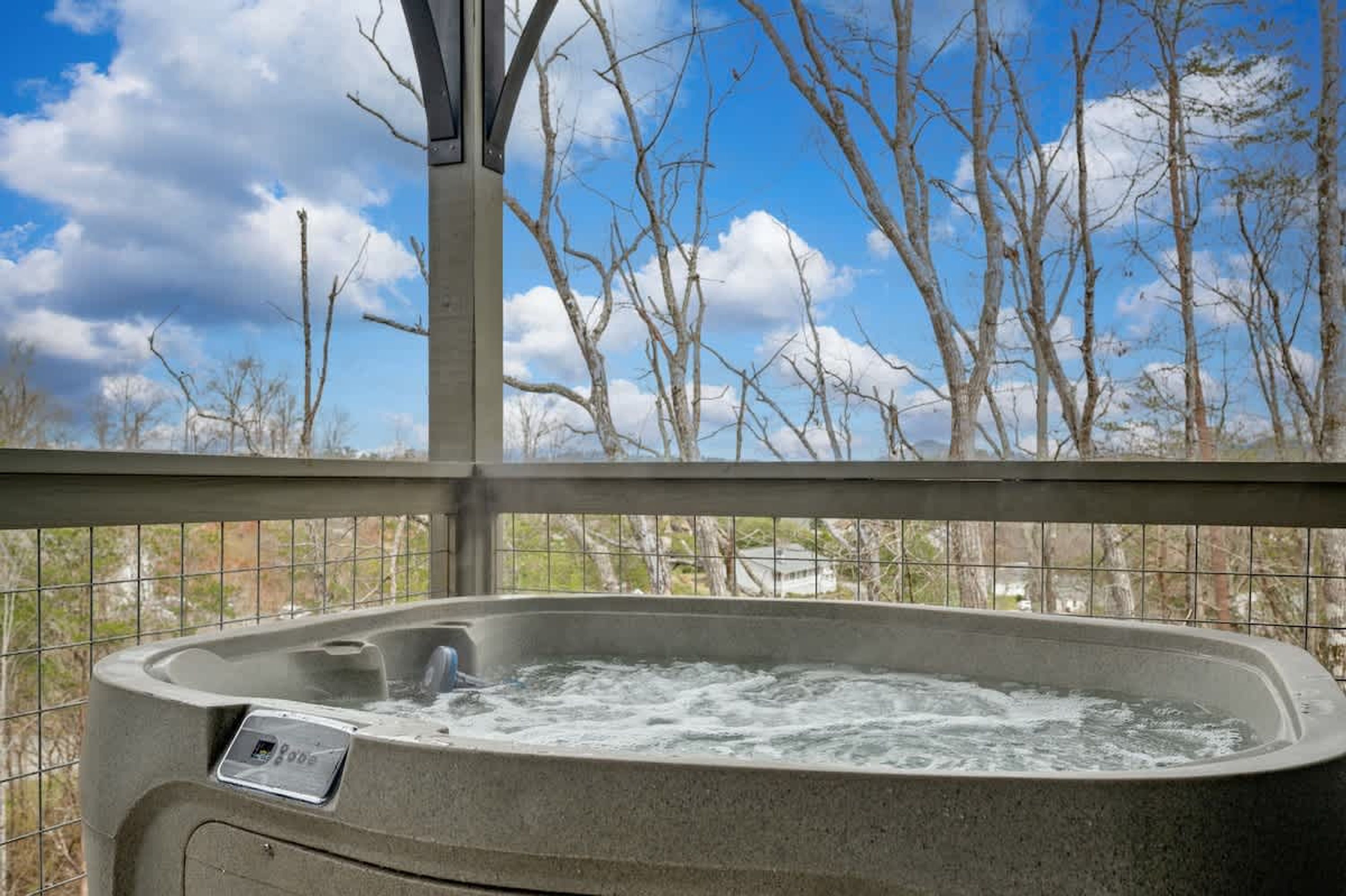 Un jacuzzi con panel de control, rodeado por una barandilla metálica, con vistas a un paisaje de árboles sin hojas y cielo despejado.