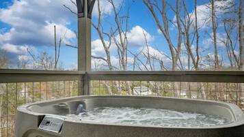 Un jacuzzi con panel de control, rodeado por una barandilla metálica, con vistas a un paisaje de árboles sin hojas y cielo despejado.