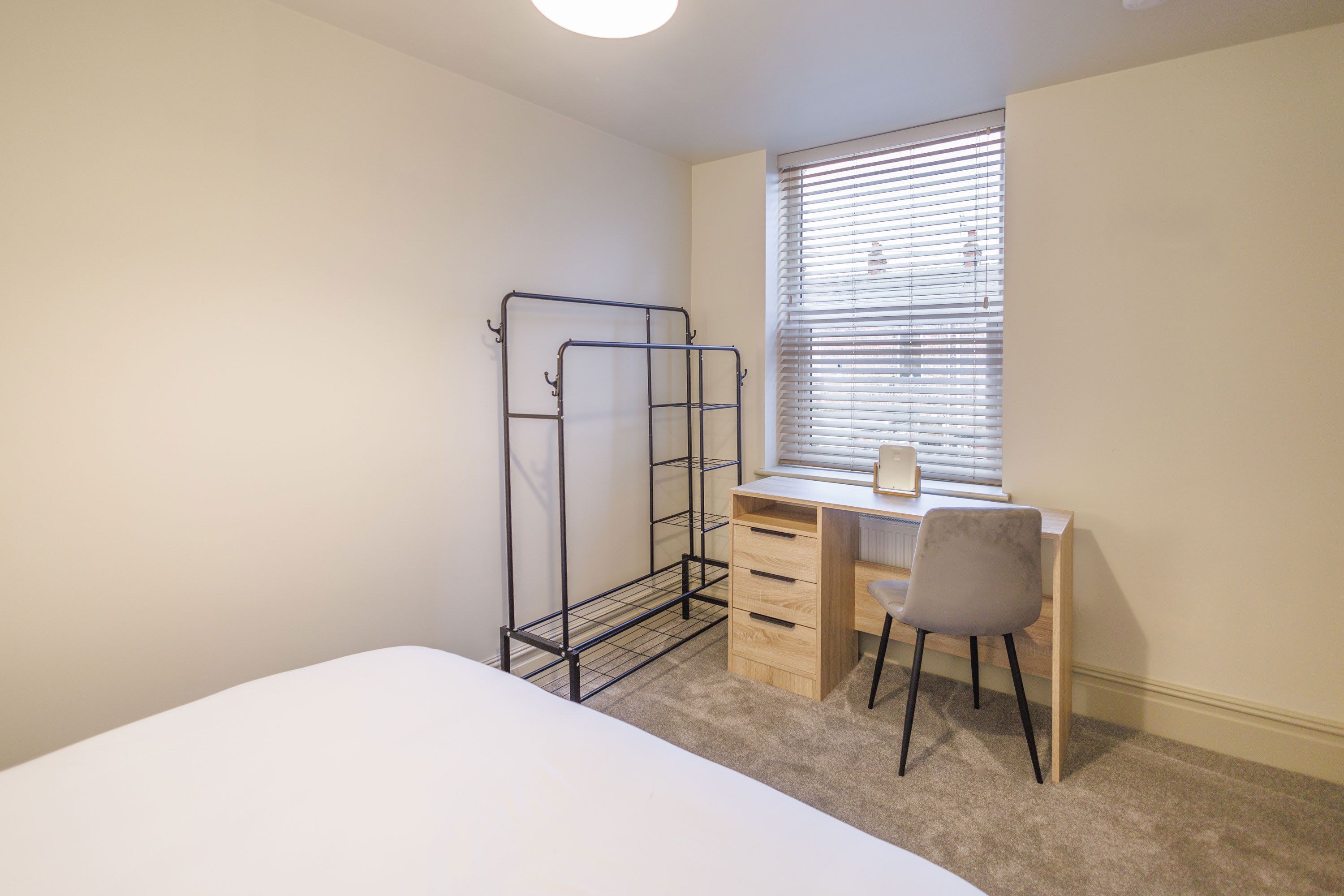A bedroom with a bed, a desk with a chair, a window with blinds, and a metal shelf unit.