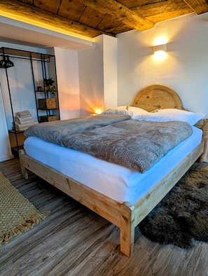 Ein hölzernes Bett mit Kopfteil, ein Nachttischchen mit gefalteten Handtüchern und eine kleine Pflanze.