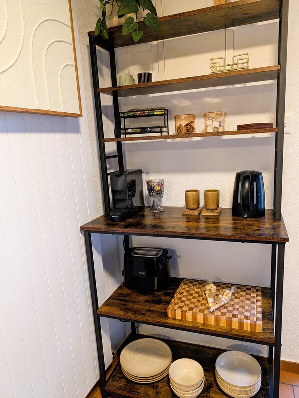 Une cuisine avec un meuble-étagère en bois, une machine à café, un grille-pain et une planche à découper à carreaux.