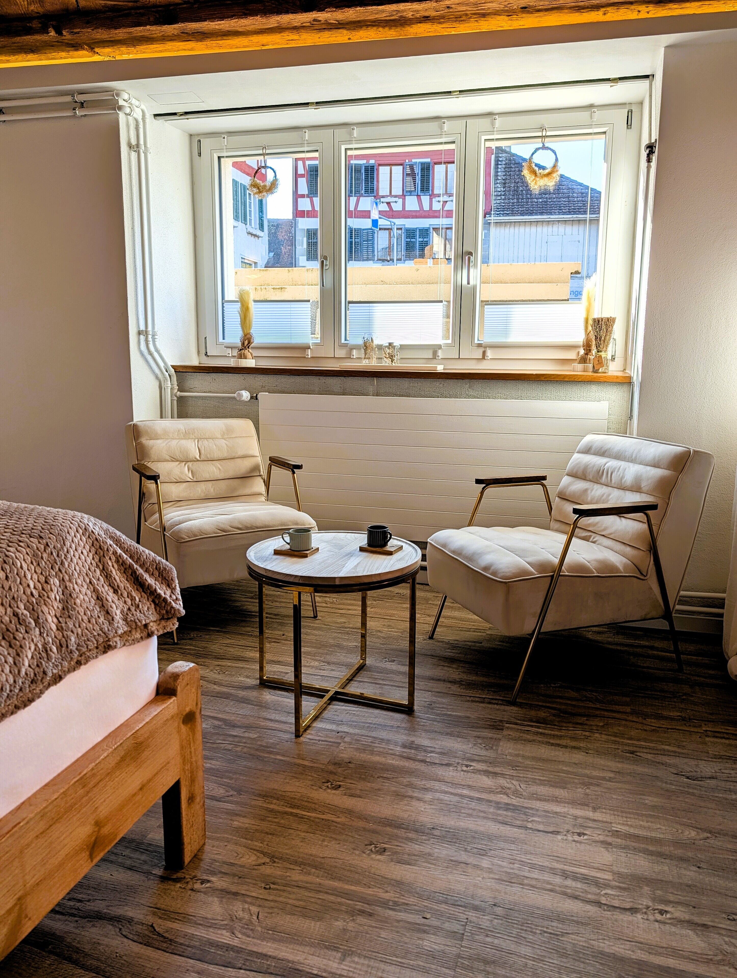 A cozy room with a wooden floor, a round table with two chairs, a bed, and a window with a view of buildings.
