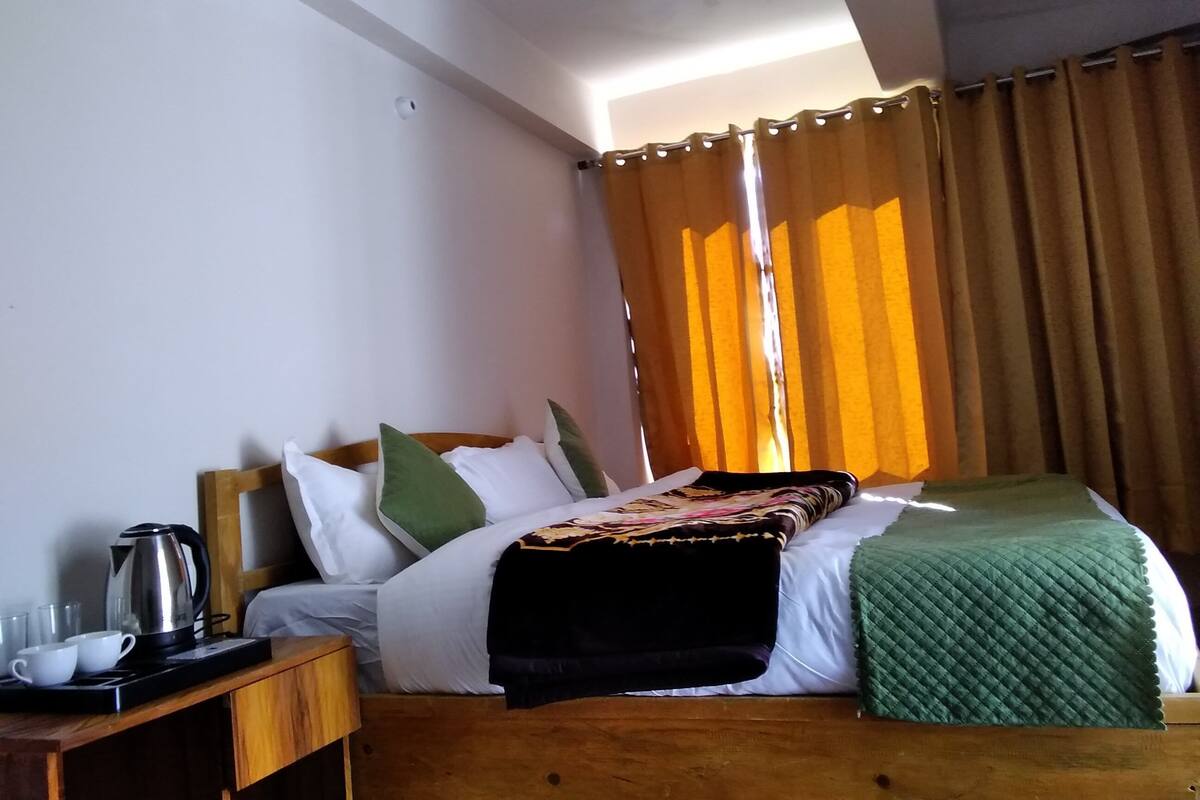 Un dormitorio con cama, almohadas, una mesita de noche con hervidor, tazas y una manta.