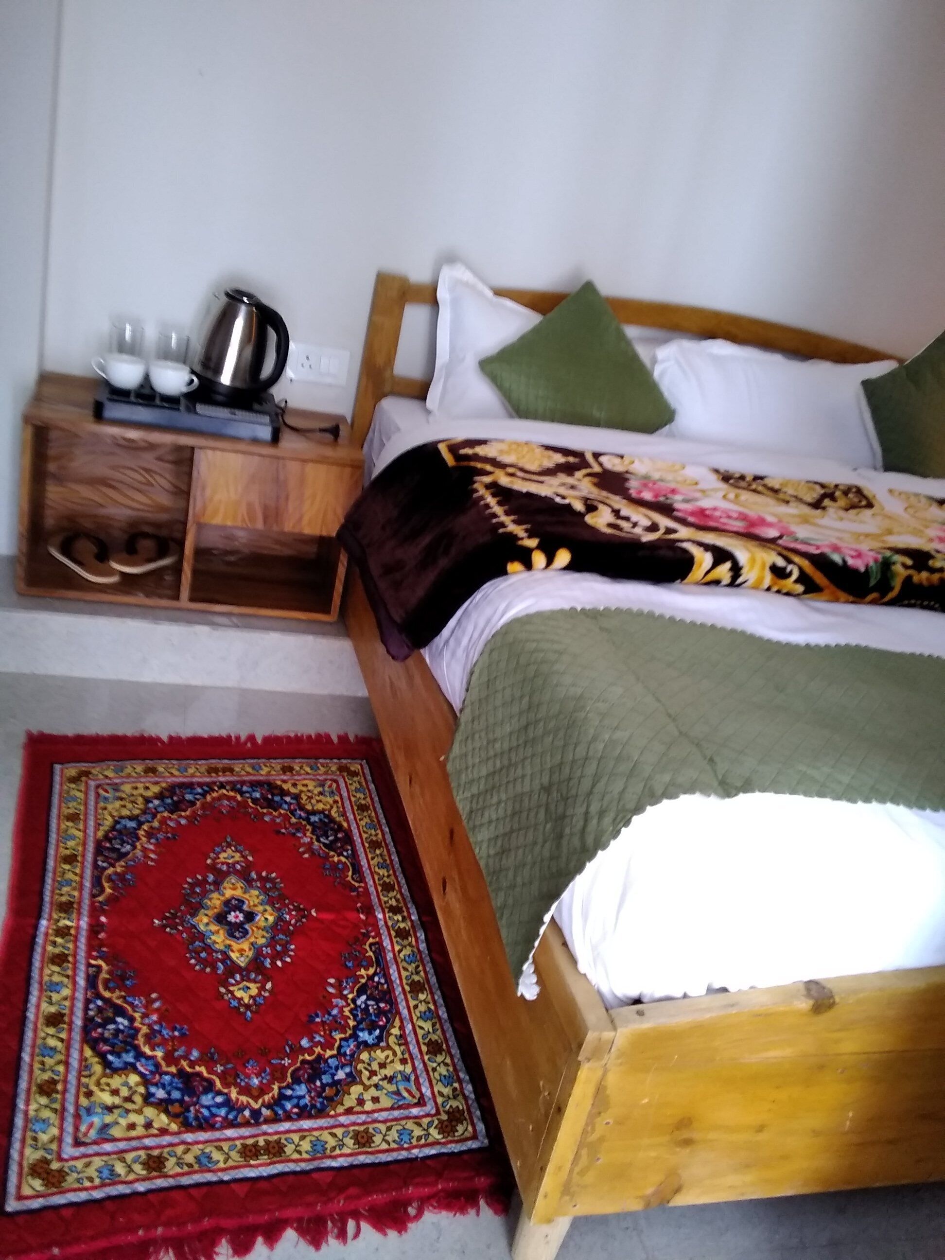 A bedroom with a wooden bed, a nightstand with a kettle, cups, and a tray, and a red patterned rug.