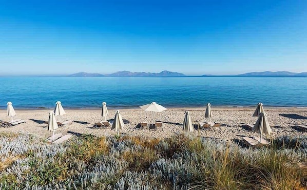 Ein Strand mit weißen Sonnenschirmen und Liegestühlen, einem klaren, blauen Himmel und entfernten Bergen.