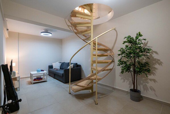 A modern living room with a spiral staircase, a grey sofa, a small white table, and a potted plant. - Home Easy Kalamata - Central Nest Private Parking (Kalamata)