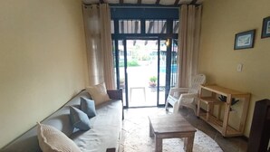 Een woonkamer met een bank, een salontafel en een houten bijzettafeltje. Er is een schuifdeur die leidt naar een buitenruimte met stoelen.