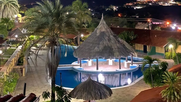 Zwembadgedeelte van een resort met een constructie met rieten dak, palmbomen en een uitzicht op verre bergen.
