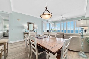A spacious living room with a wooden dining table, white chairs, a beige sofa, and a ceiling fan. - Regency Isle 1102  3 3  2 covered parking spots  PENTHOUSE (Orange Beach)