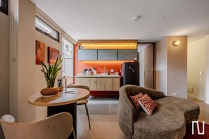 Une cuisine moderne avec des meubles en bois, un îlot central et un réfrigérateur. On y trouve un coin repas avec une table ronde et deux chaises. Une plante en pot est posée sur la table, et des œuvres d’art ornent le mur.