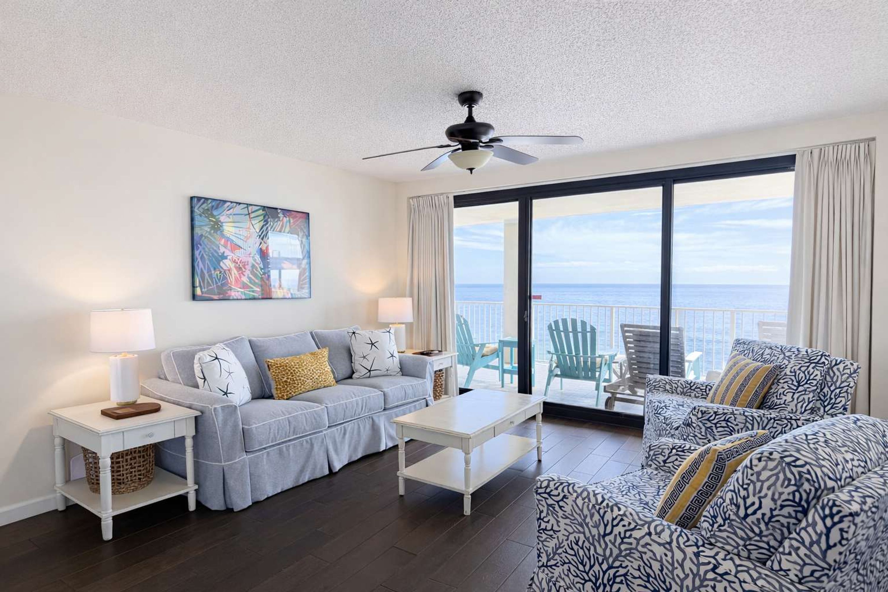 A living room with a sofa, a coffee table, a ceiling fan, a painting, and a view of the ocean.