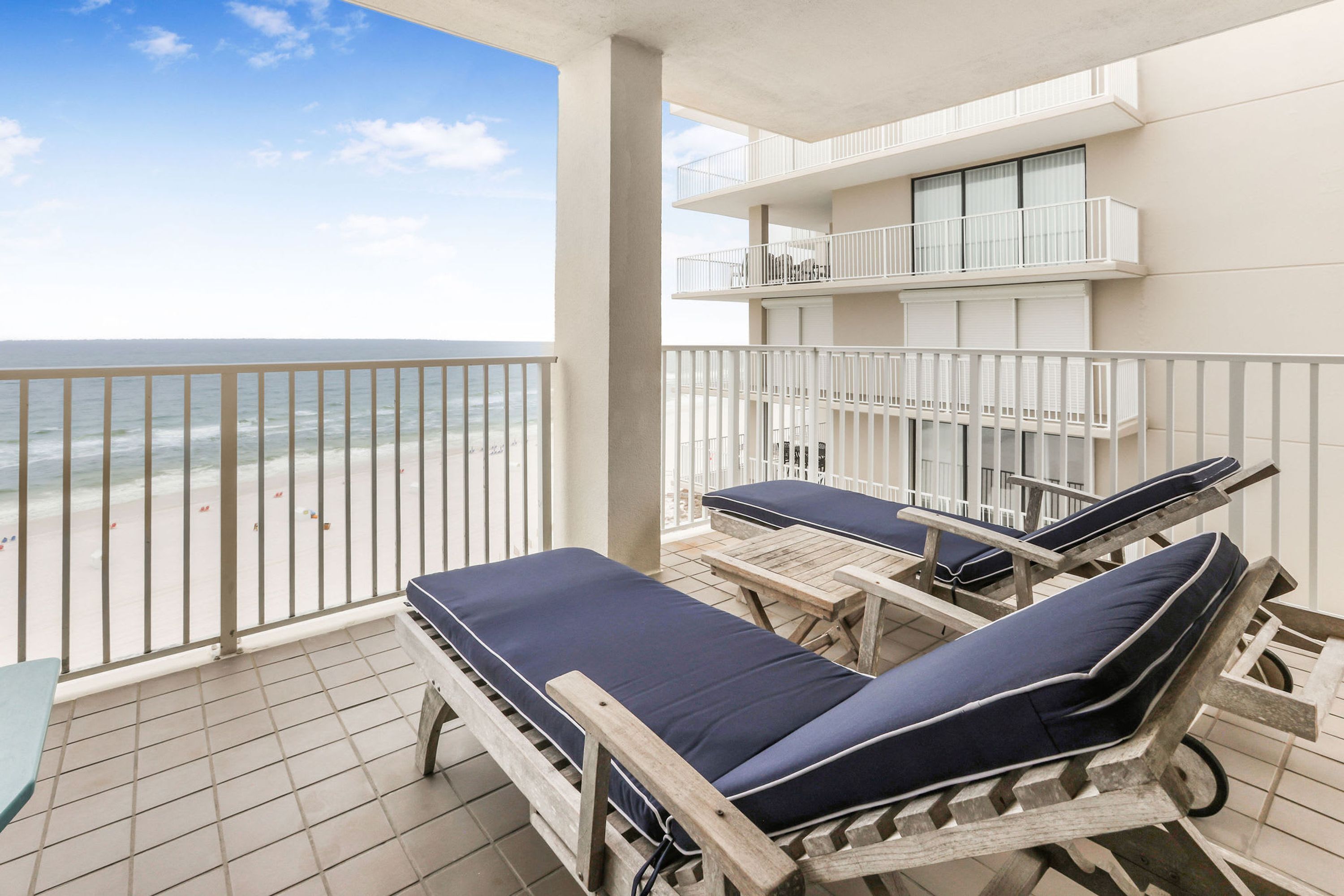 A balcony with two lounge chairs and a table, overlooking a beach.