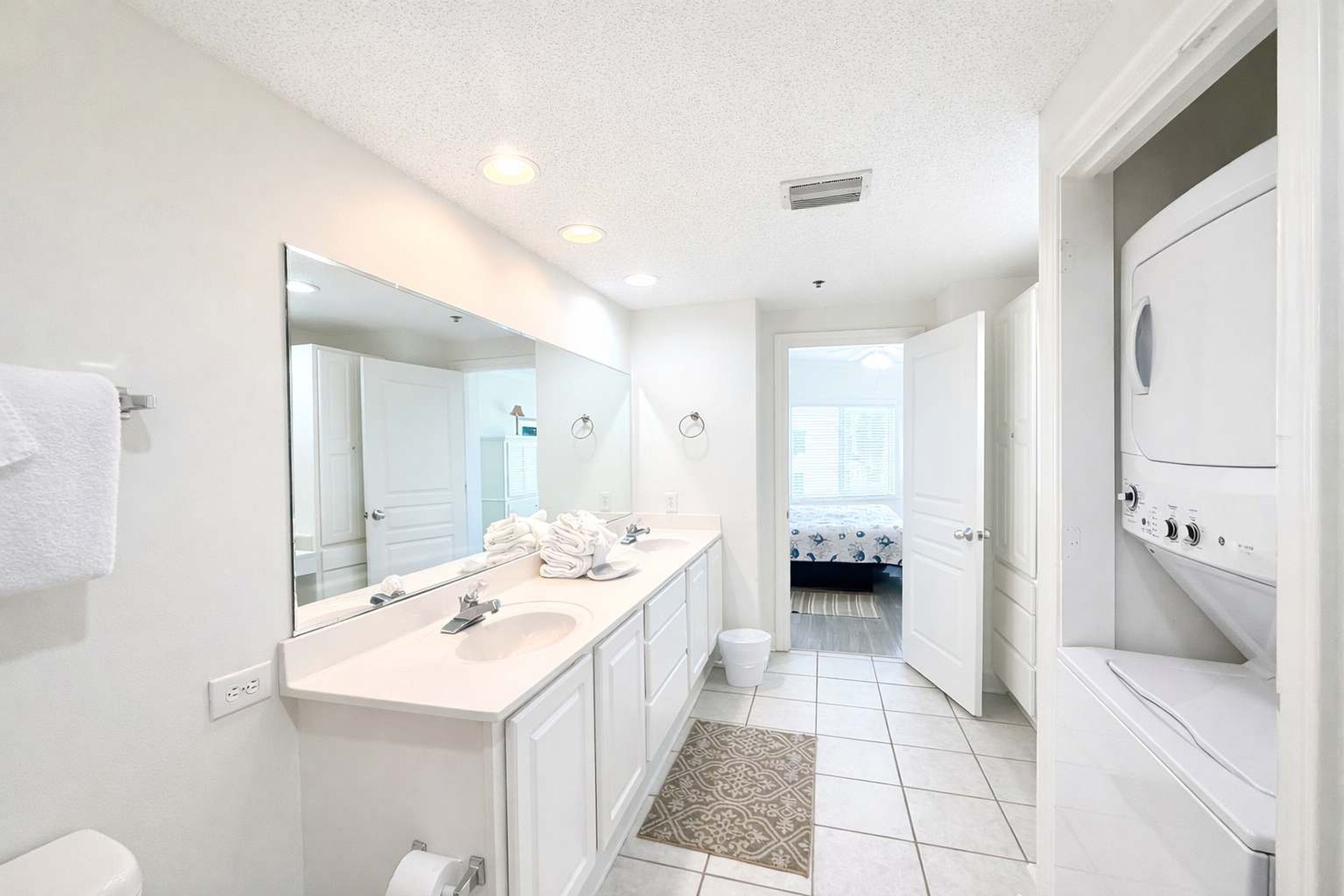 A modern bathroom with a large mirror, dual sinks, a toilet, and a built-in washer-dryer unit.