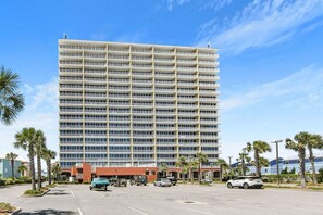 Un hôtel à plusieurs étages, doté d’une façade uniforme, un parking où sont garées plusieurs voitures, et des palmiers tout autour.