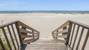 Une promenade en bois mène à une plage de sable fin, sous un ciel bleu et dégagé.