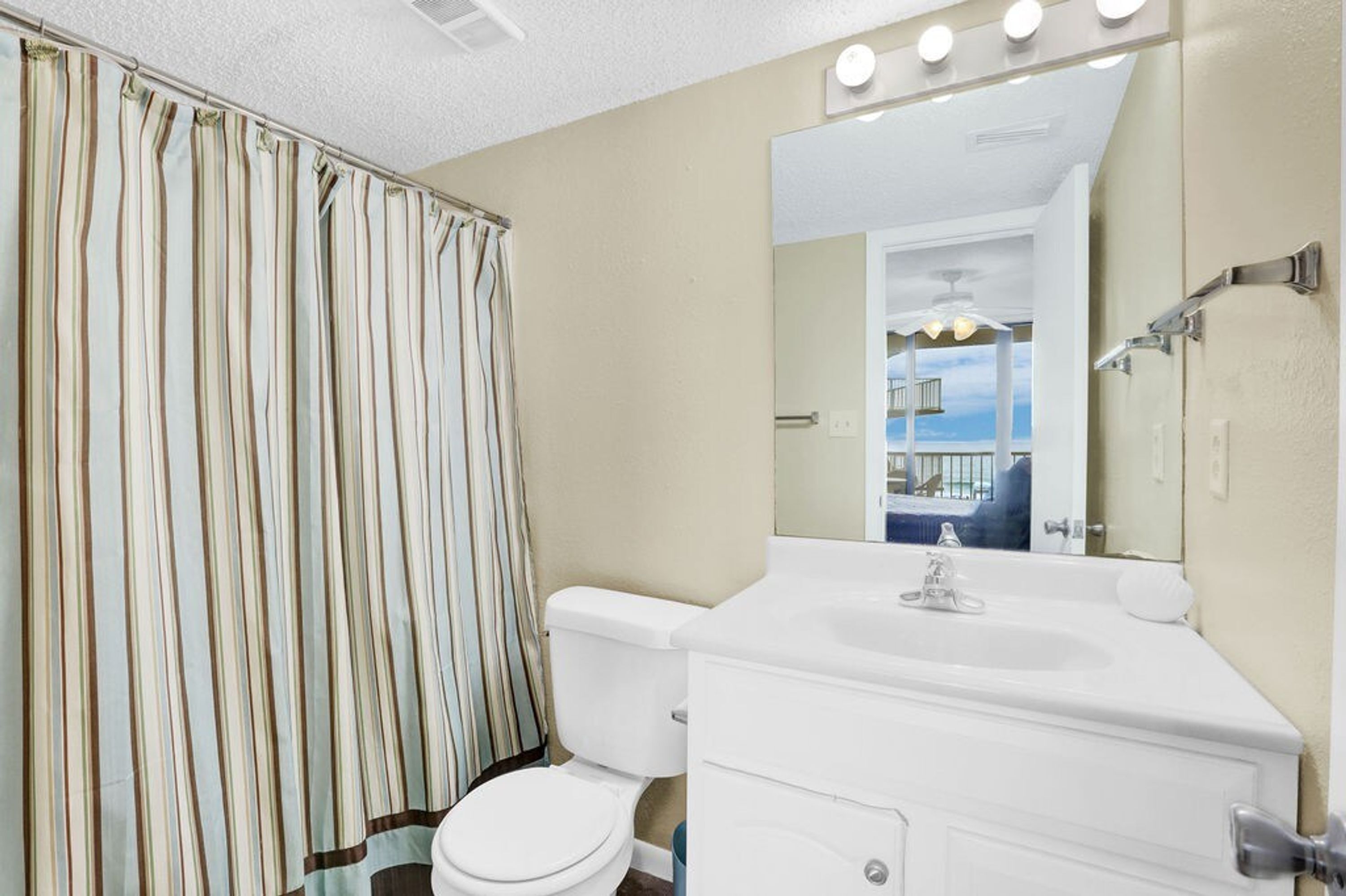Un baño con cortina de ducha a rayas, un inodoro blanco, un lavabo blanco y un espejo con luces arriba.