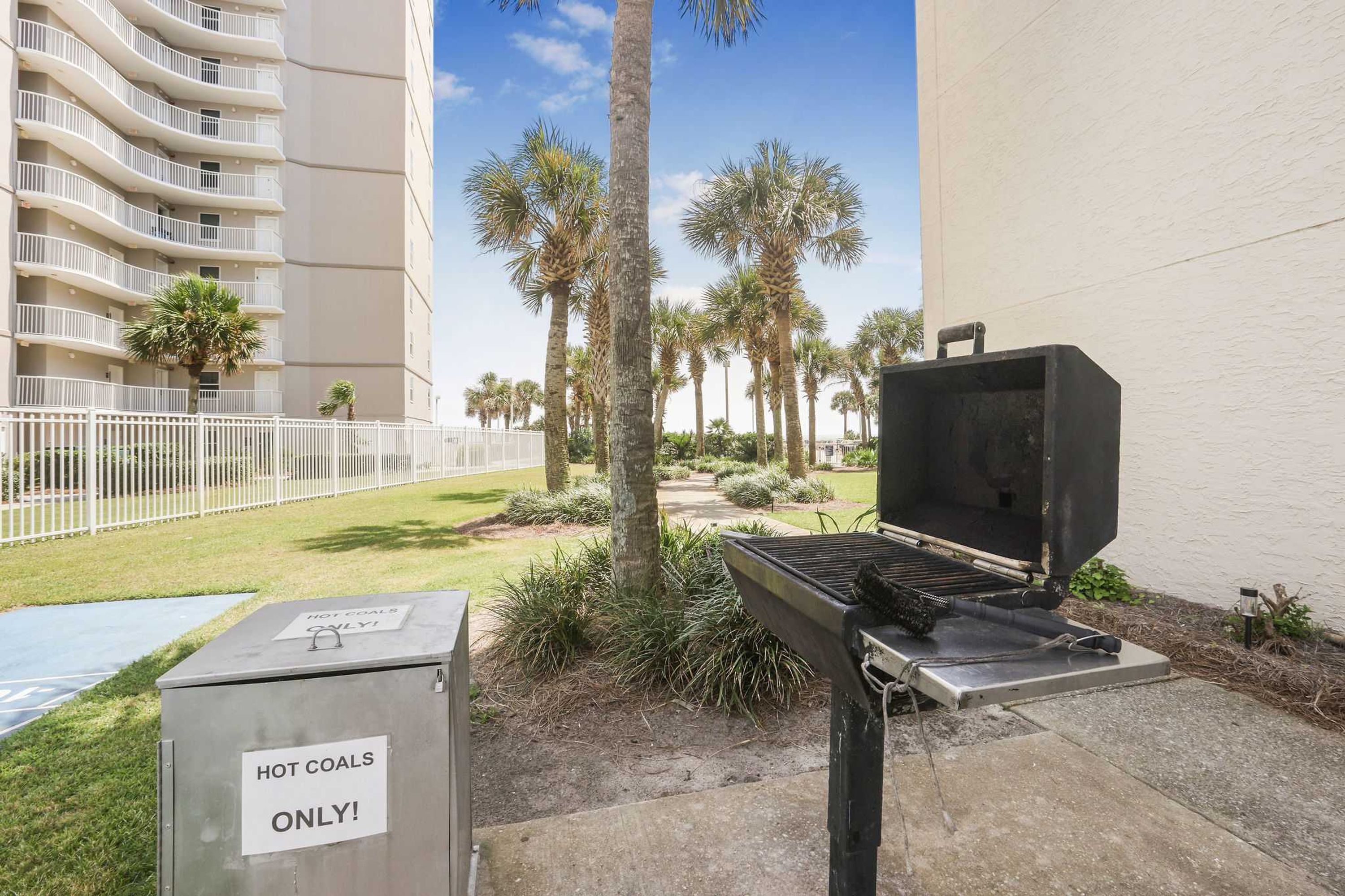 A barbecue grill with a sign indicating it's for hot coals only, set against a backdrop of a tall building and palm trees.