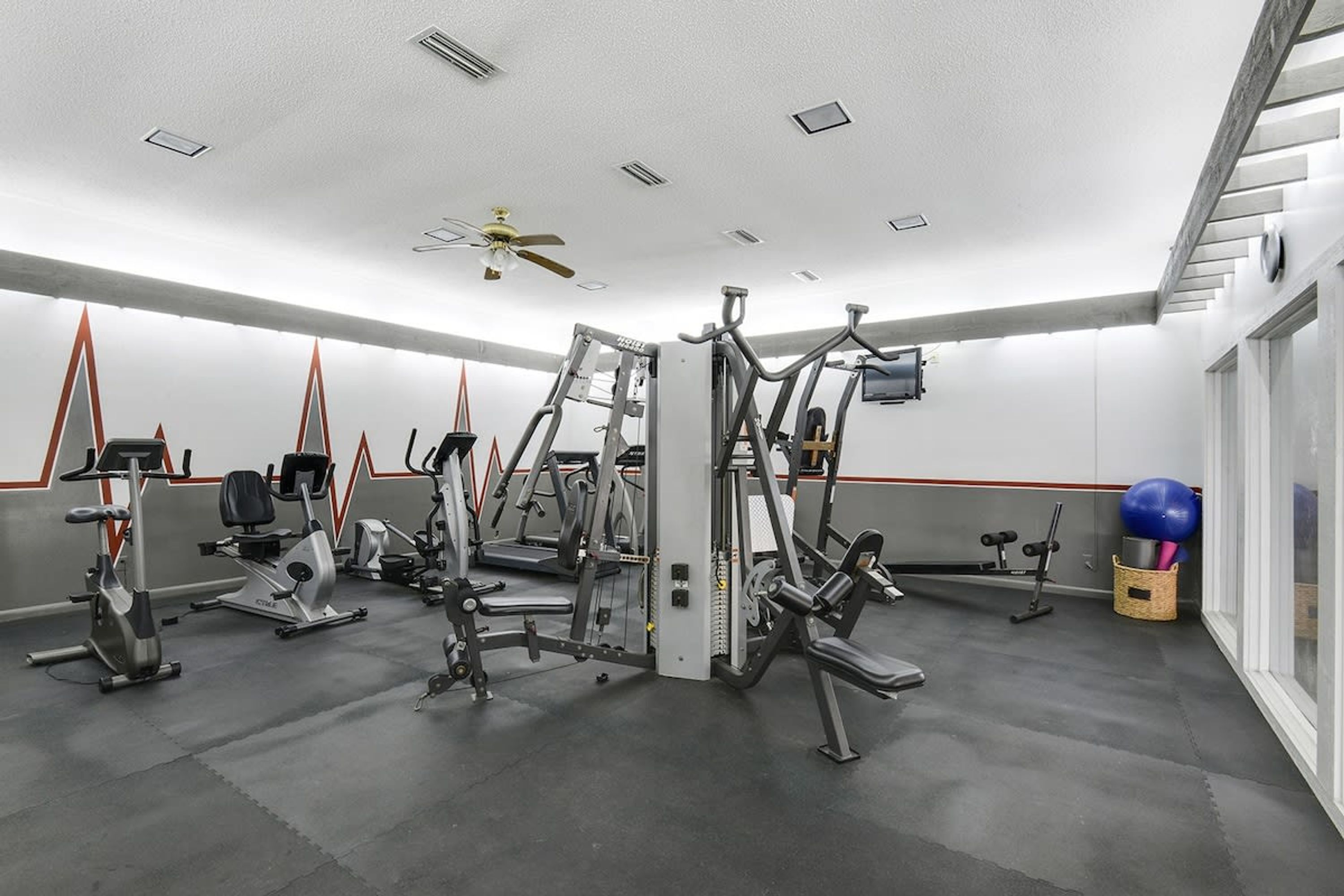 Un gimnasio con diversas máquinas de ejercicio, un ventilador de techo y un amplio ventanal a la derecha.
