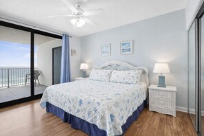Une chambre à coucher avec un lit, deux tables de chevet avec des lampes, un ventilateur de plafond et une vue sur l’océan par une porte coulissante.