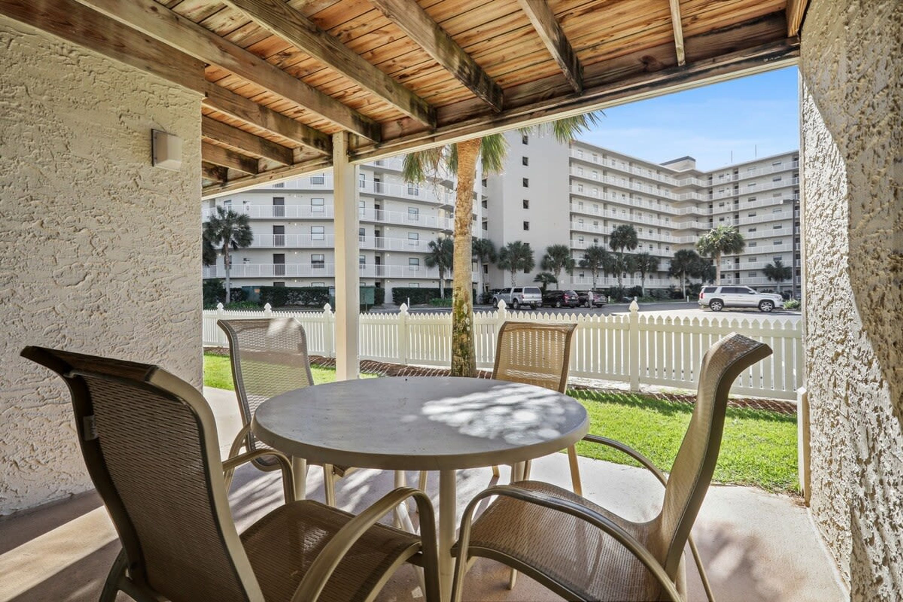 A covered outdoor seating area with a round table and four chairs, overlooking a street with parked cars and a white picket fence.