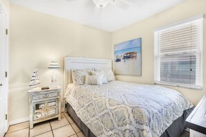 Quarto com cama, mesa de cabeceira, abajur, pintura e janela com persianas.
