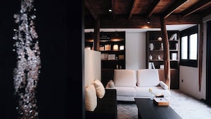 A modern living room with a white sofa, a black ottoman, and a wooden coffee table. There is a bookshelf filled with books and decorative items. The room has a wooden ceiling and large windows. - Sanfuyizhai Homestay (Taizhou)