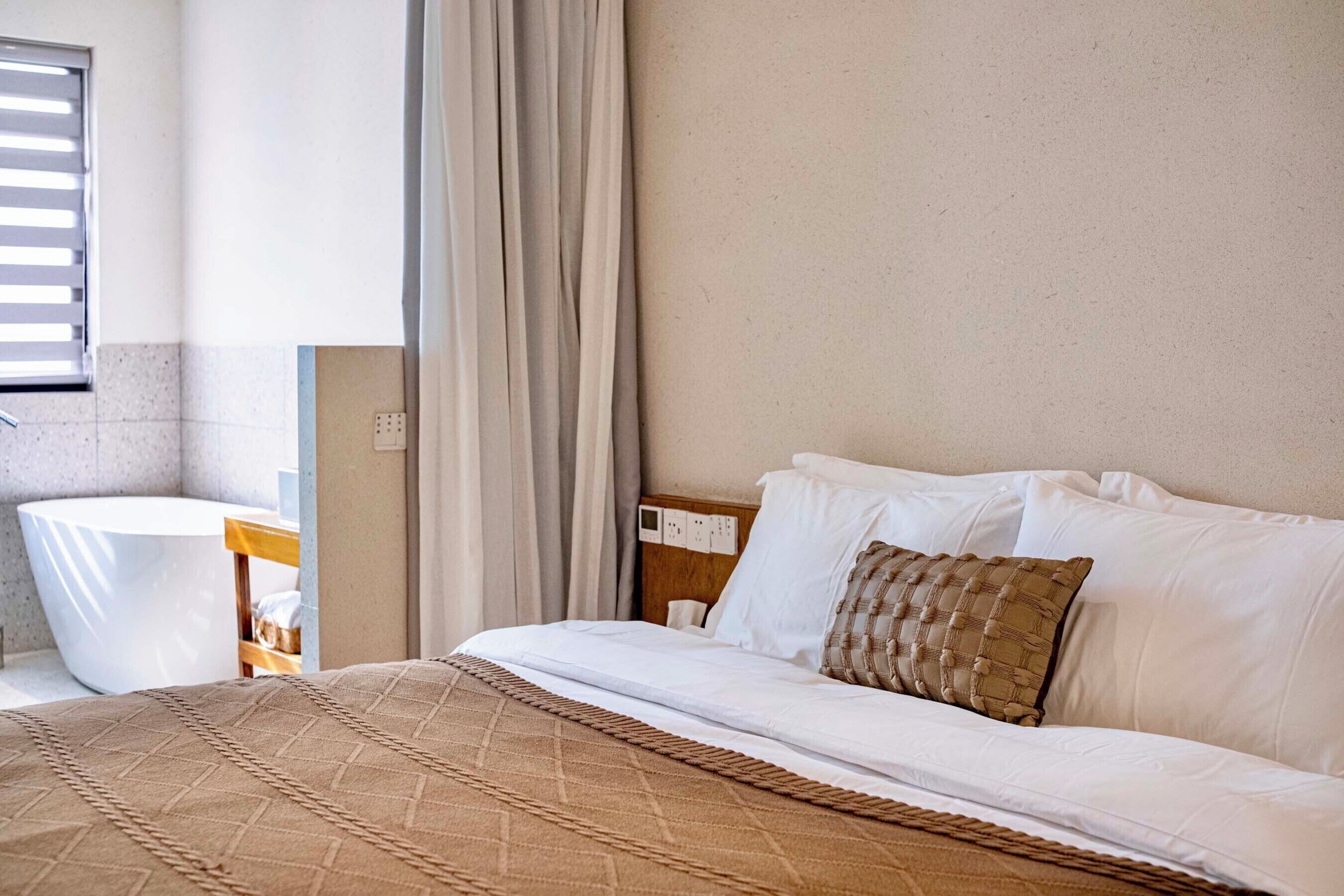 A hotel room with a bed, a bedside table, a white chair, and a window with blinds.