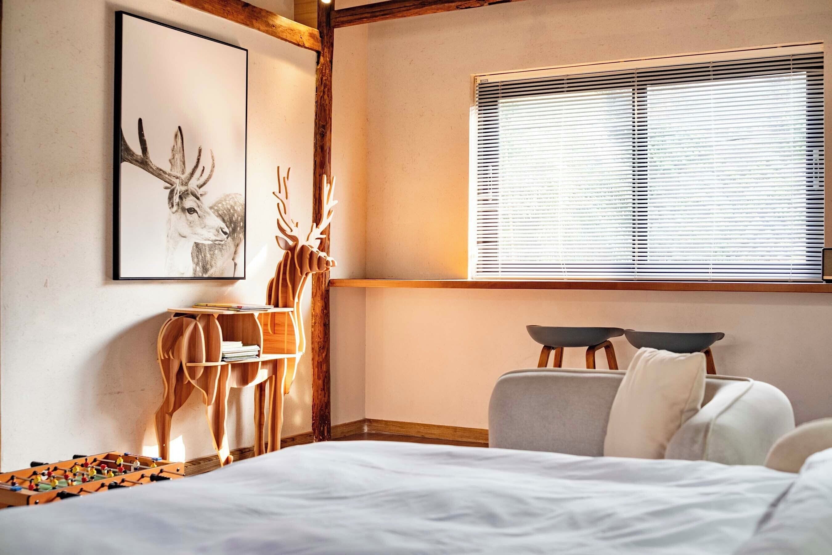 A bedroom with a bed, a desk with a chair, a window with blinds, and a wall-mounted picture of a deer.