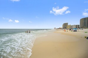 Ein Strand mit sandigem Ufer, Meereswellen und einigen Personen. Im Hintergrund sind Gebäude zu sehen.