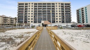 A multi-story hotel with a central entrance, a sandy beach in the foreground, and a wooden boardwalk leading to the entrance. - Island Wind West 177  Next to Hangout  pool (Gulf Shores)