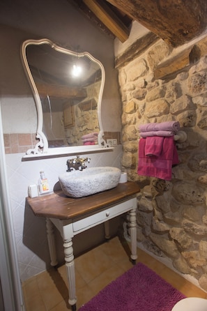 A bathroom with a stone wall, a wooden vanity with a white sink, a mirror, and a purple rug.