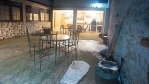 A stone-walled outdoor area with a table and chairs, a stone fireplace, and a well-lit interior visible through an open door. - CASA RURAL INMA (Muñoveros)