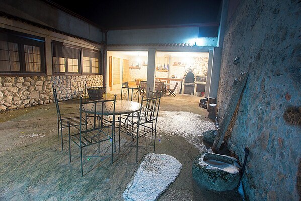 A stone-walled outdoor area with a table and chairs, a stone fireplace, and a well-lit interior visible through an open door.