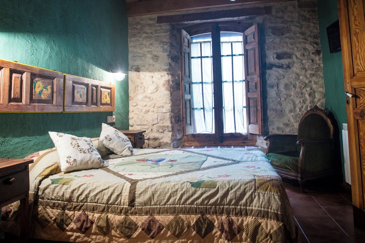 A rustic bedroom with a wooden ceiling, a stone wall, a bed with a quilt, and a window with open shutters.