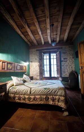 Une chambre rustique avec un plafond en bois, un mur en pierre, un lit recouvert d’une courtepointe et une fenêtre aux volets ouverts.
