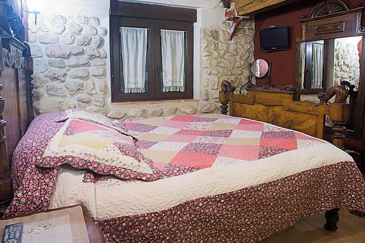 A bedroom with a wooden ceiling, stone wall, a bed with a floral quilt, a wooden dresser, and a stone fireplace.